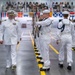 A formal U.S. Navy military ceremony taking place in a large indoor arena or hangar. A senior officer in dress whites with gold insignia walks past a rifle honor guard also dressed in white uniforms. A large audience of spectators and seated sailors fills the background, with international and naval signal flags decorating the walls.