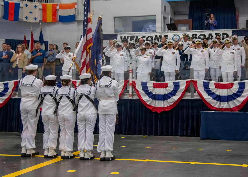 A formal U.S. Navy commissioning or change of command ceremony is taking place indoors, likely in a hangar bay or large military facility. A color guard in enlisted dress whites presents the American flag and Navy flag while officers in dress whites salute from a raised stage. A 'Welcome Aboard' banner is displayed prominently behind the stage, which is decorated with patriotic red, white, and blue bunting.