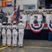 A formal U.S. Navy commissioning or change of command ceremony is taking place indoors, likely in a hangar bay or large military facility. A color guard in enlisted dress whites presents the American flag and Navy flag while officers in dress whites salute from a raised stage. A 'Welcome Aboard' banner is displayed prominently behind the stage, which is decorated with patriotic red, white, and blue bunting.