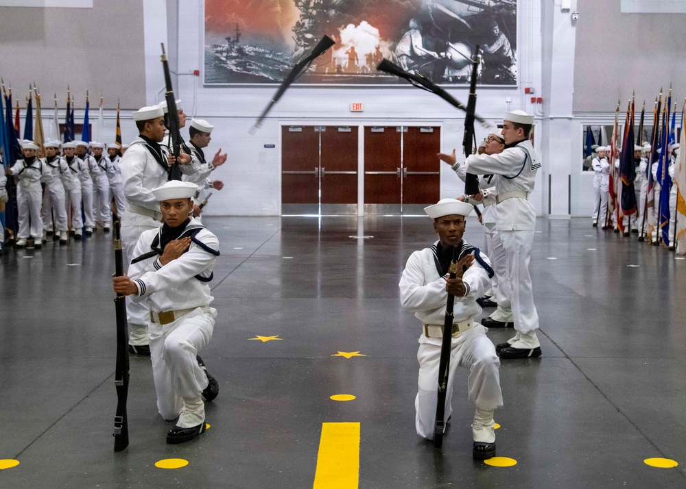 A U.S. Navy drill team performs a precision rifle exhibition inside a large ceremonial hall, with four performers in the foreground executing synchronized kneeling and rifle-tossing maneuvers. The performers wear traditional Navy dress whites with white Dixie cup covers, black shoes, and white belts. In the background, rows of sailors in formation stand alongside color guard members holding flags, in front of a large naval battle mural.