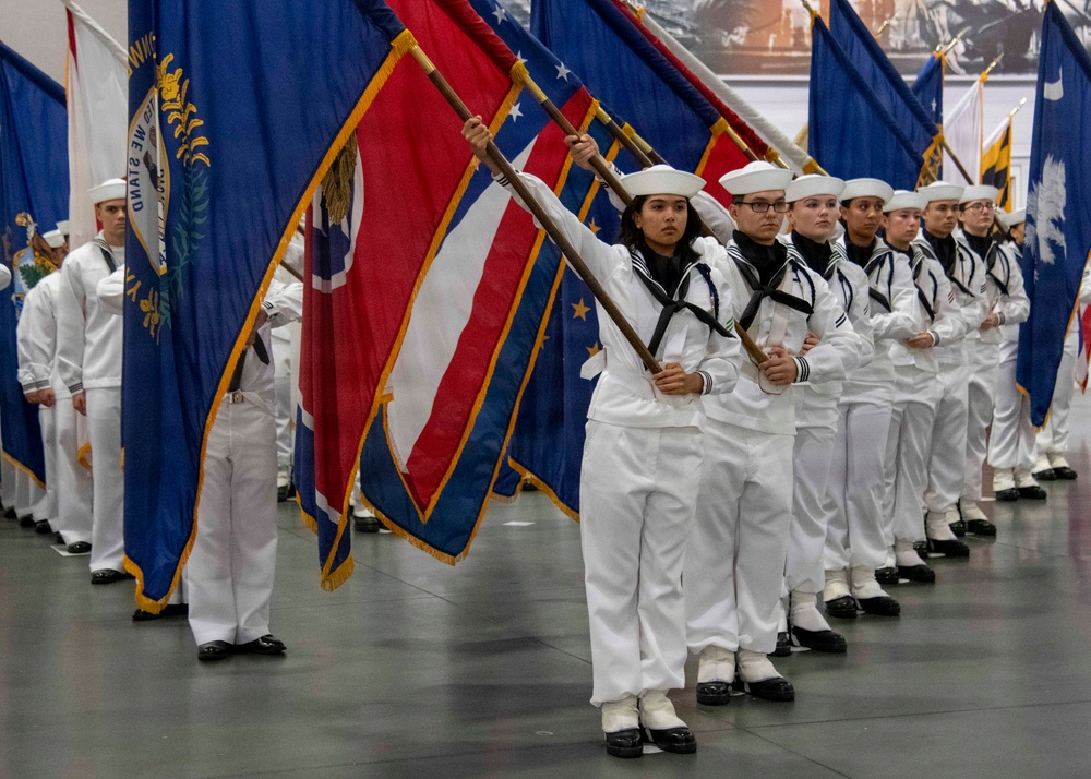 A formation of U.S. Navy sailors in dress white uniforms participates in a formal color guard ceremony, carrying multiple state and organizational flags. The personnel are arranged in a precise diagonal line inside a large indoor venue, suggesting a graduation or official military ceremony. Both male and female sailors are visible, all wearing the distinctive white Crackerjack-style Navy dress uniforms with white dixie cup covers.