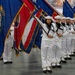 A formation of U.S. Navy sailors in dress white uniforms participates in a formal color guard ceremony, carrying multiple state and organizational flags. The personnel are arranged in a precise diagonal line inside a large indoor venue, suggesting a graduation or official military ceremony. Both male and female sailors are visible, all wearing the distinctive white Crackerjack-style Navy dress uniforms with white dixie cup covers.
