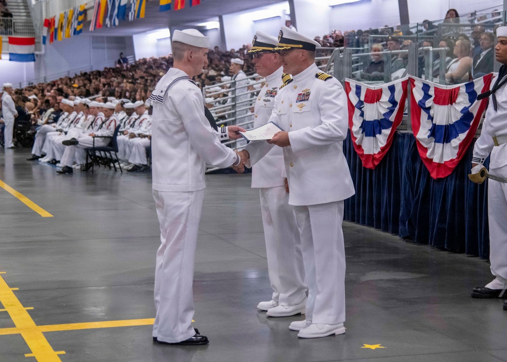 A formal U.S. Navy graduation or awards ceremony taking place in a large indoor facility, likely a drill hall or hangar. A junior enlisted sailor in dress white uniform is receiving a document or certificate from a senior officer, while another officer stands nearby. The venue is filled with seated sailors and civilian spectators in the background.