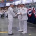 A formal U.S. Navy graduation or awards ceremony taking place in a large indoor facility, likely a drill hall or hangar. A junior enlisted sailor in dress white uniform is receiving a document or certificate from a senior officer, while another officer stands nearby. The venue is filled with seated sailors and civilian spectators in the background.