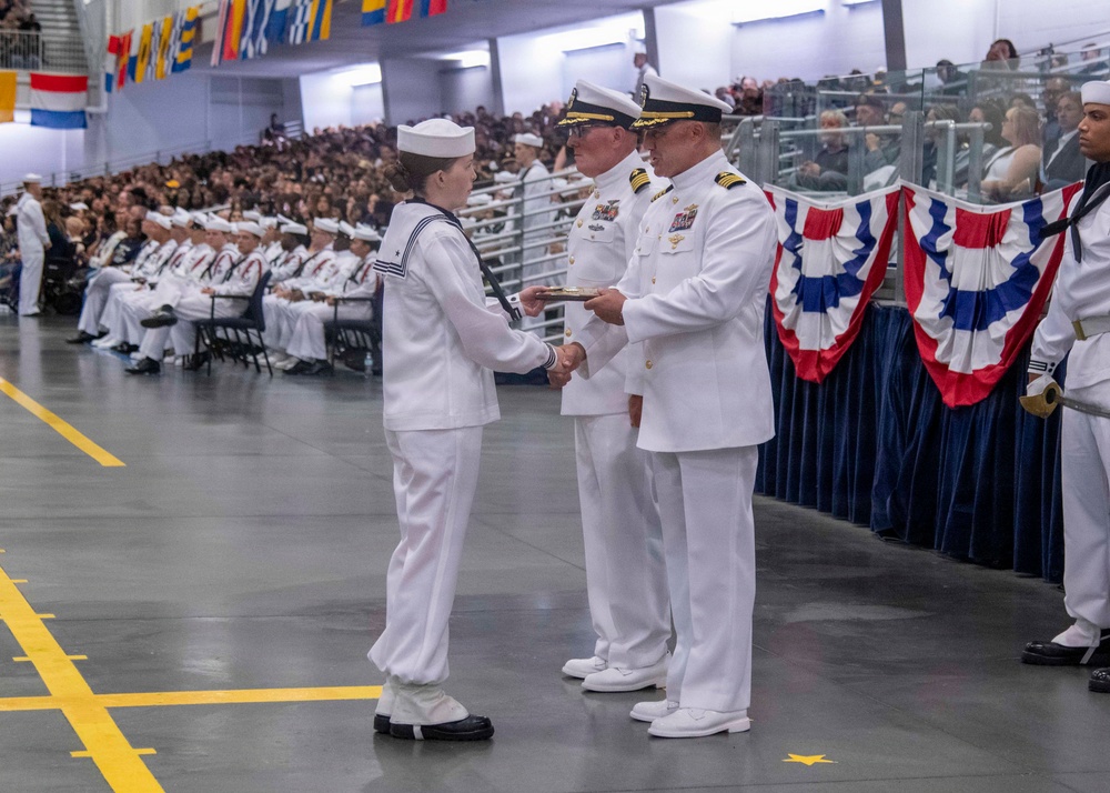 A U.S. Navy graduation or pass-in-review ceremony is taking place in a large indoor venue. A junior enlisted sailor in dress white uniform is receiving what appears to be a certificate or award from two senior officers wearing dress white uniforms with gold insignia. The venue is decorated with red, white, and blue bunting, international signal flags, and is filled with seated sailors and civilian spectators.
