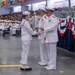 A U.S. Navy graduation or pass-in-review ceremony is taking place in a large indoor venue. A junior enlisted sailor in dress white uniform is receiving what appears to be a certificate or award from two senior officers wearing dress white uniforms with gold insignia. The venue is decorated with red, white, and blue bunting, international signal flags, and is filled with seated sailors and civilian spectators.