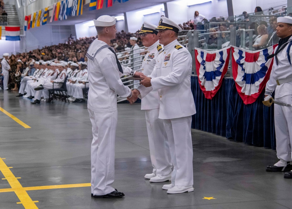 A formal U.S. Navy graduation or commissioning ceremony taking place in a large indoor venue. A senior naval officer in dress whites is shaking hands with an enlisted sailor, likely presenting a certificate or diploma. The event is attended by a large audience of family members and fellow sailors seated in the background.