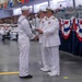 A formal U.S. Navy graduation or commissioning ceremony taking place in a large indoor venue. A senior naval officer in dress whites is shaking hands with an enlisted sailor, likely presenting a certificate or diploma. The event is attended by a large audience of family members and fellow sailors seated in the background.