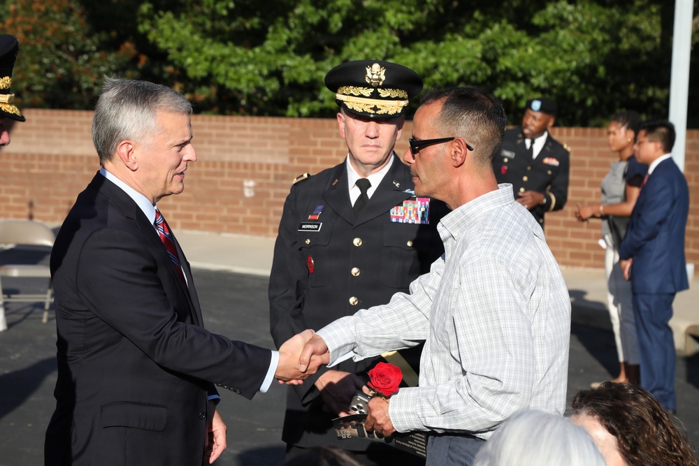 A formal outdoor ceremony appears to be taking place, likely a military or civic recognition event. A civilian in a dark suit is shaking hands with another civilian holding a red rose and what appears to be a plaque or award. A U.S. Army officer in dress uniform with the name tag 'Morrison' stands between them, observing the exchange.