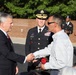 A formal outdoor ceremony appears to be taking place, likely a military or civic recognition event. A civilian in a dark suit is shaking hands with another civilian holding a red rose and what appears to be a plaque or award. A U.S. Army officer in dress uniform with the name tag 'Morrison' stands between them, observing the exchange.