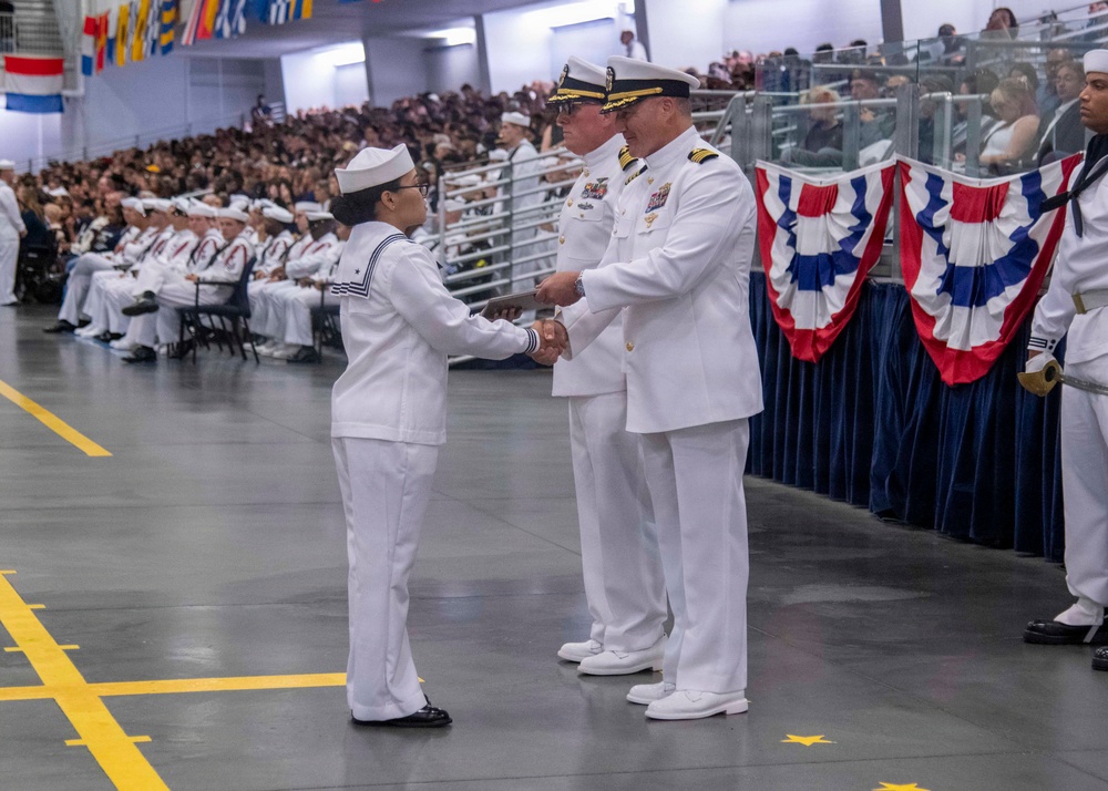 A U.S. Navy graduation or pass-in-review ceremony is taking place in a large indoor venue. A junior enlisted sailor is shaking hands with a senior officer while receiving what appears to be a certificate or document. Rows of seated sailors in white dress uniforms and a large audience of civilians are visible in the background.