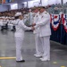 A U.S. Navy graduation or pass-in-review ceremony is taking place in a large indoor venue. A junior enlisted sailor is shaking hands with a senior officer while receiving what appears to be a certificate or document. Rows of seated sailors in white dress uniforms and a large audience of civilians are visible in the background.