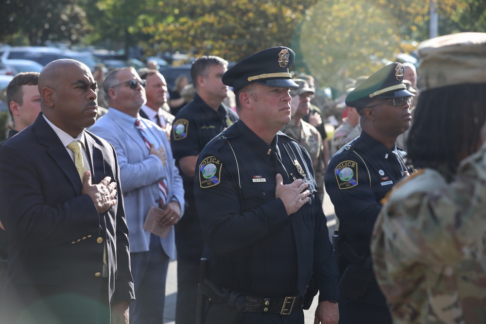 A formal outdoor ceremony featuring law enforcement officers and military personnel standing at attention with hands over their hearts, likely during a Pledge of Allegiance or National Anthem. Officers wearing 'State Capitol NC' police patches are prominently visible alongside a civilian in a suit and military personnel in camouflage uniforms. The gathering appears to be a commemorative or official public ceremony.