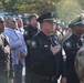 A formal outdoor ceremony featuring law enforcement officers and military personnel standing at attention with hands over their hearts, likely during a Pledge of Allegiance or National Anthem. Officers wearing 'State Capitol NC' police patches are prominently visible alongside a civilian in a suit and military personnel in camouflage uniforms. The gathering appears to be a commemorative or official public ceremony.