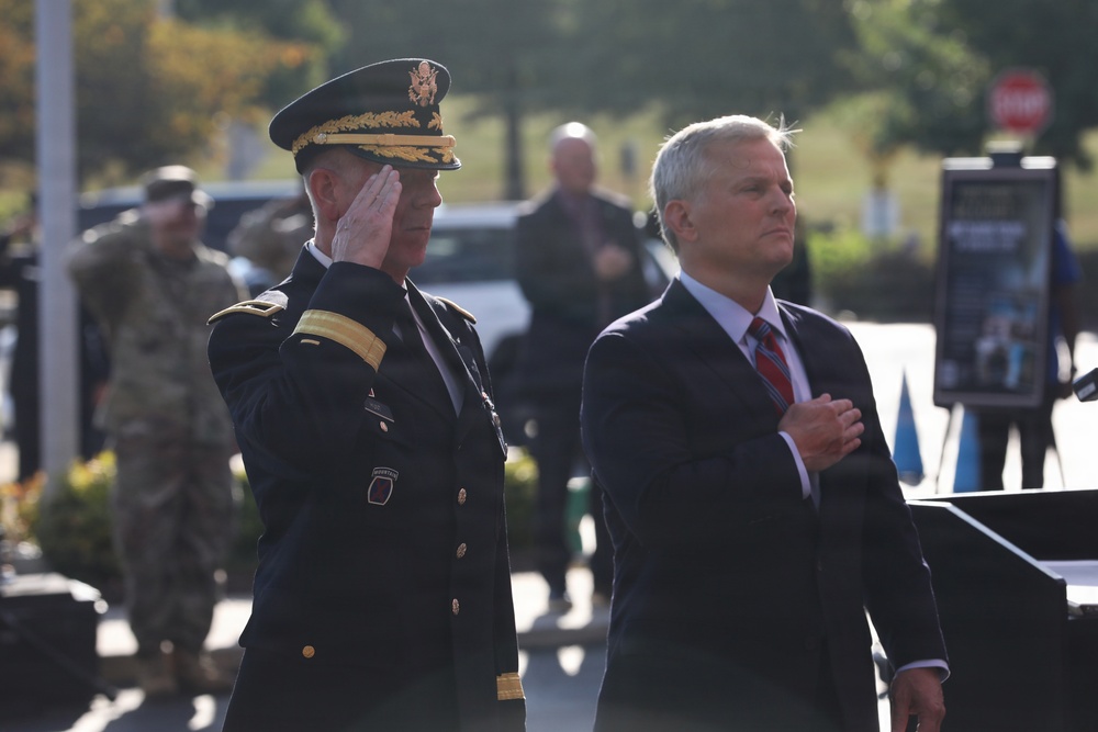 Two men stand at attention during what appears to be a formal military ceremony. A uniformed U.S. Army general officer renders a salute while a civilian in a dark suit places his right hand over his heart, likely during the playing of the national anthem or a similar ceremonial moment. The outdoor setting and background personnel suggest an official public military event.