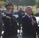 Two men stand at attention during what appears to be a formal military ceremony. A uniformed U.S. Army general officer renders a salute while a civilian in a dark suit places his right hand over his heart, likely during the playing of the national anthem or a similar ceremonial moment. The outdoor setting and background personnel suggest an official public military event.