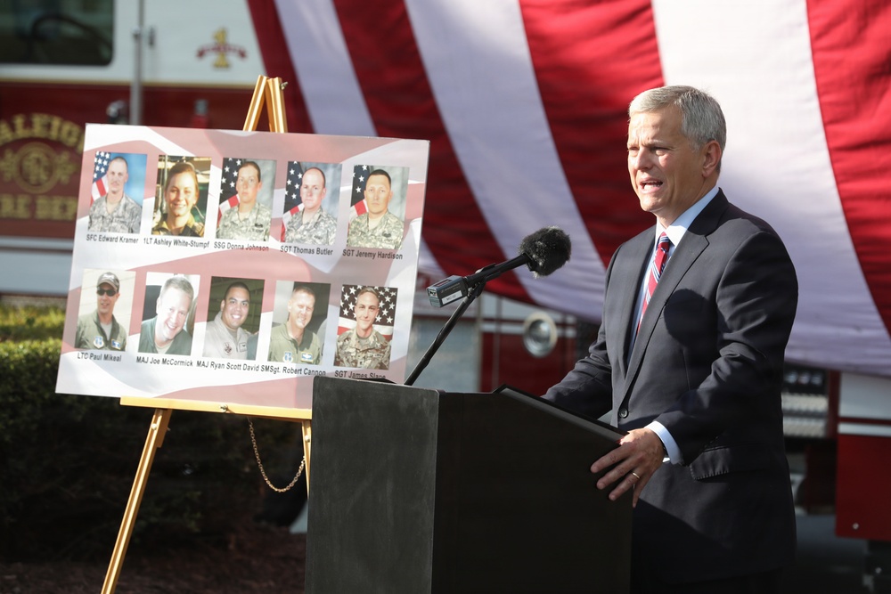 A man in a dark suit speaks at a podium with a microphone during what appears to be a memorial or tribute ceremony. Behind him is a display board featuring photographs and names of military personnel, along with a large American flag backdrop. The setting appears to be outdoors, possibly at a fire department facility based on visible signage.