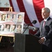 A man in a dark suit speaks at a podium with a microphone during what appears to be a memorial or tribute ceremony. Behind him is a display board featuring photographs and names of military personnel, along with a large American flag backdrop. The setting appears to be outdoors, possibly at a fire department facility based on visible signage.