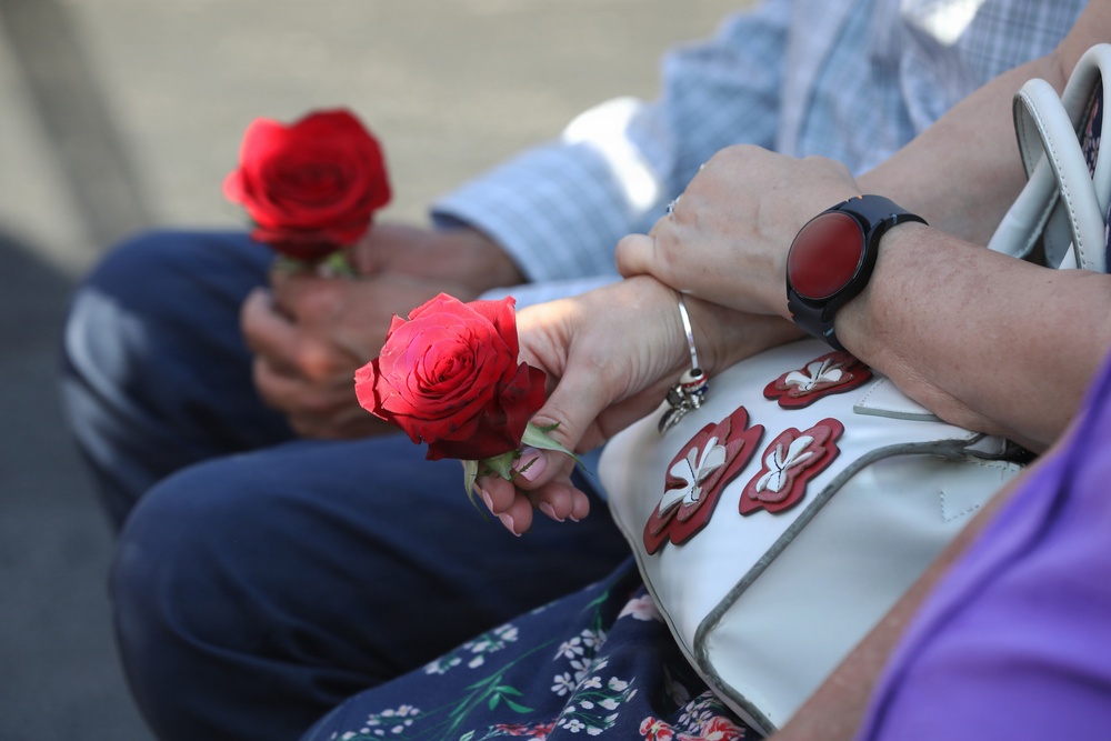 Two people are seated side by side, each holding a single red rose. One person is carrying a white handbag with red floral appliqué decorations and wearing a dark smartwatch with a red face. The scene suggests a formal or commemorative occasion where roses have been distributed.