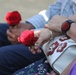 Two people are seated side by side, each holding a single red rose. One person is carrying a white handbag with red floral appliqué decorations and wearing a dark smartwatch with a red face. The scene suggests a formal or commemorative occasion where roses have been distributed.