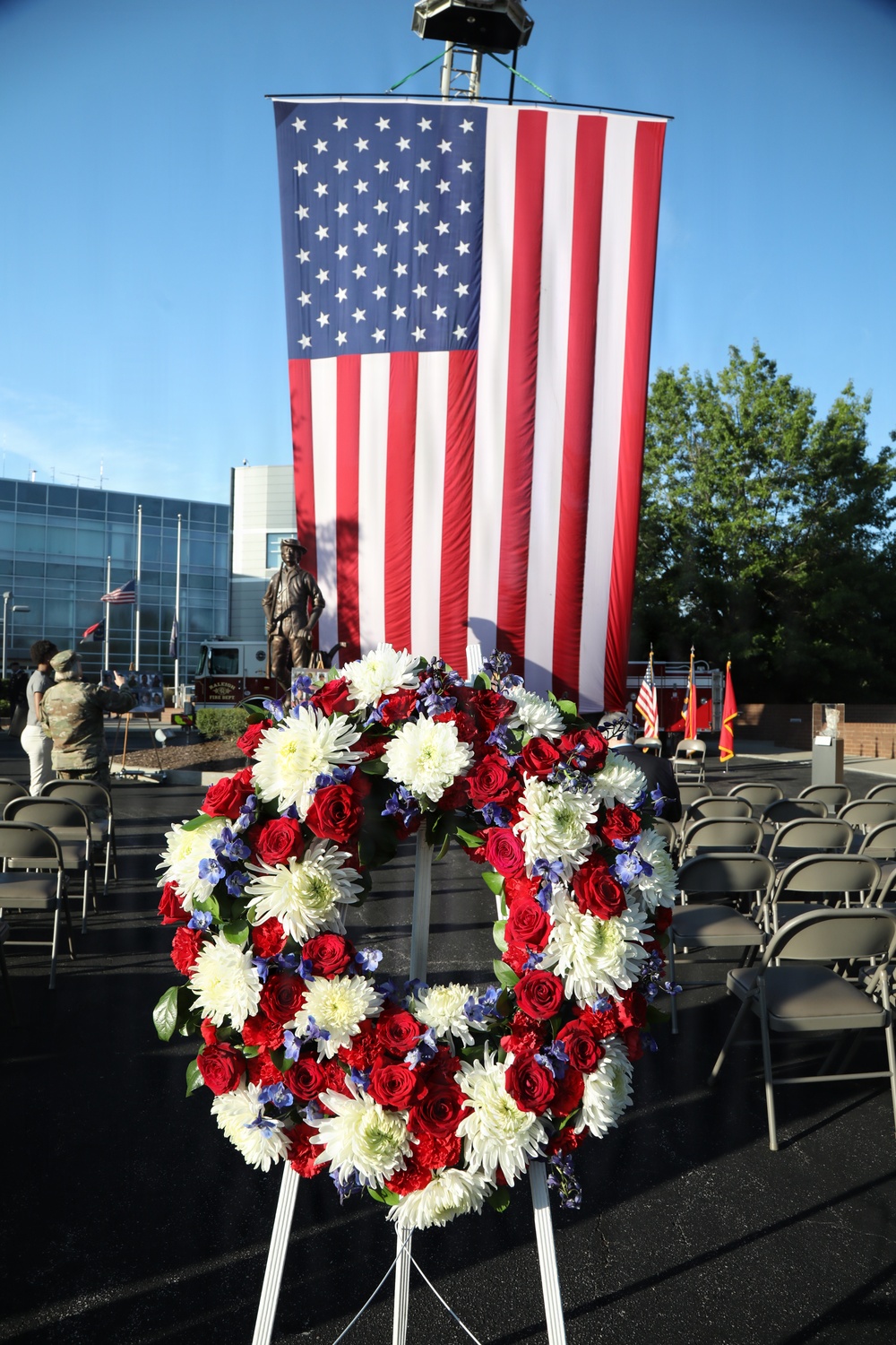 A patriotic memorial ceremony is taking place outdoors, featuring a large vertically-hung American flag as a backdrop. In the foreground, a floral wreath composed of red roses, white chrysanthemums, and blue flowers is displayed on a white easel stand. The setting includes rows of folding chairs, a bronze statue of a firefighter, a fire truck, and military/organizational flags, suggesting a first responder memorial event.