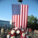A patriotic memorial ceremony is taking place outdoors, featuring a large vertically-hung American flag as a backdrop. In the foreground, a floral wreath composed of red roses, white chrysanthemums, and blue flowers is displayed on a white easel stand. The setting includes rows of folding chairs, a bronze statue of a firefighter, a fire truck, and military/organizational flags, suggesting a first responder memorial event.
