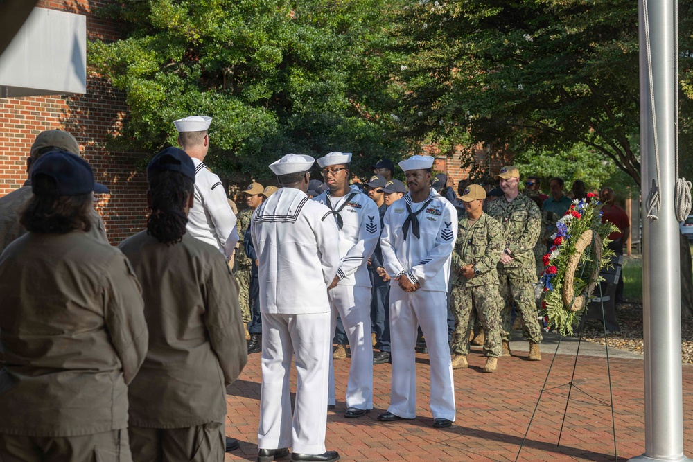 A military ceremony is taking place outdoors on a brick plaza, featuring U.S. Navy sailors in white dress uniforms and personnel in camouflage uniforms. A patriotic wreath decorated with red, white, and blue flowers is visible on the right side, suggesting a memorial or commemorative event. Civilian observers are present in the foreground watching the proceedings.