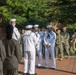 A military ceremony is taking place outdoors on a brick plaza, featuring U.S. Navy sailors in white dress uniforms and personnel in camouflage uniforms. A patriotic wreath decorated with red, white, and blue flowers is visible on the right side, suggesting a memorial or commemorative event. Civilian observers are present in the foreground watching the proceedings.