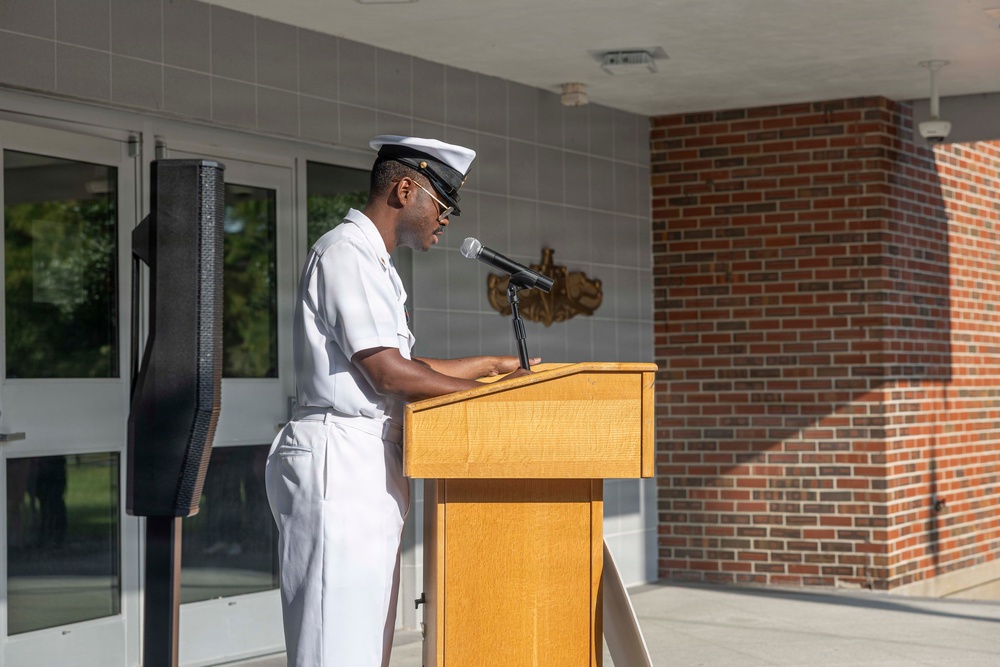 A U.S. Navy enlisted sailor in white dress uniform is speaking at a wooden podium equipped with a microphone during what appears to be an official military ceremony or event. The setting is an outdoor or semi-covered area in front of a building with brick and tile facade. A military emblem or insignia is visible mounted on the wall behind the podium.