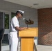 A U.S. Navy enlisted sailor in white dress uniform is speaking at a wooden podium equipped with a microphone during what appears to be an official military ceremony or event. The setting is an outdoor or semi-covered area in front of a building with brick and tile facade. A military emblem or insignia is visible mounted on the wall behind the podium.