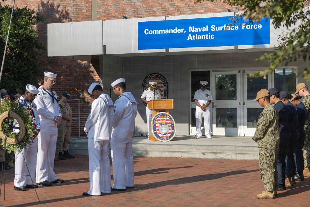 A military ceremony is taking place outside the headquarters of Commander, Naval Surface Force Atlantic. Personnel in various Navy uniforms are assembled in formation while an officer speaks at a podium bearing the Commander Naval Surface Force Atlantic seal. A memorial wreath is visible on the left side, suggesting this is a commemorative or memorial event.