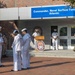 A military ceremony is taking place outside the headquarters of Commander, Naval Surface Force Atlantic. Personnel in various Navy uniforms are assembled in formation while an officer speaks at a podium bearing the Commander Naval Surface Force Atlantic seal. A memorial wreath is visible on the left side, suggesting this is a commemorative or memorial event.