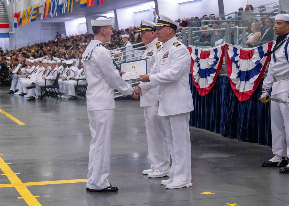 A U.S. Navy graduation or awards ceremony taking place in a large indoor venue, likely a drill hall or hangar. A junior enlisted sailor in dress whites is receiving a certificate or diploma from a senior officer, also in dress whites, while another officer stands behind. A large audience of sailors and civilians fills the bleachers in the background.