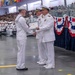 A U.S. Navy graduation or awards ceremony taking place in a large indoor venue, likely a drill hall or hangar. A junior enlisted sailor in dress whites is receiving a certificate or diploma from a senior officer, also in dress whites, while another officer stands behind. A large audience of sailors and civilians fills the bleachers in the background.