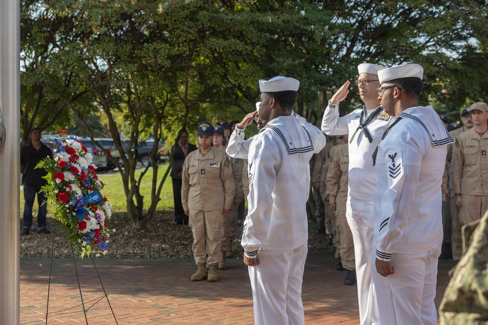 Three U.S. Navy sailors in dress white uniforms are rendering a salute beside a red, white, and blue memorial wreath during what appears to be a formal military ceremony. A formation of sailors and personnel in tan/khaki working uniforms stands at attention in the background. The setting is an outdoor brick plaza surrounded by trees, suggesting a commemorative or memorial event.