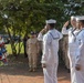 Three U.S. Navy sailors in dress white uniforms are rendering a salute beside a red, white, and blue memorial wreath during what appears to be a formal military ceremony. A formation of sailors and personnel in tan/khaki working uniforms stands at attention in the background. The setting is an outdoor brick plaza surrounded by trees, suggesting a commemorative or memorial event.