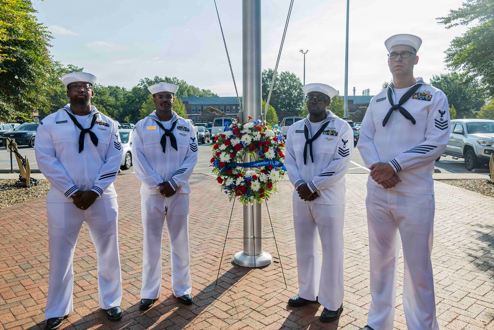 Four U.S. Navy sailors in dress white uniforms stand at attention flanking a memorial wreath mounted on a stand. The wreath features red, white, and blue flowers with a ribbon reading 'September 11' visible on it. The ceremony appears to be a 9/11 commemorative wreath-laying event at a military installation.