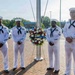 Four U.S. Navy sailors in dress white uniforms stand at attention flanking a memorial wreath mounted on a stand. The wreath features red, white, and blue flowers with a ribbon reading 'September 11' visible on it. The ceremony appears to be a 9/11 commemorative wreath-laying event at a military installation.