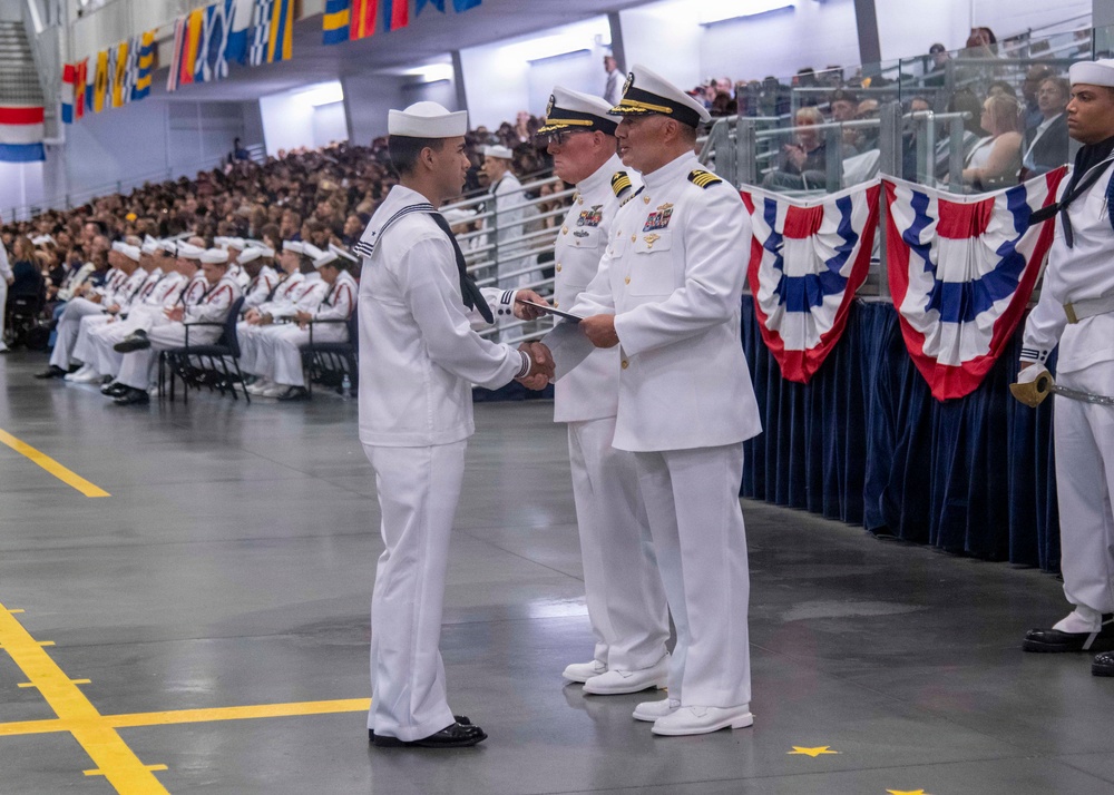 A formal U.S. Navy graduation or awards ceremony taking place in a large indoor facility. A sailor in dress whites is receiving a certificate or document with a handshake from a senior officer, while another officer stands nearby. A large audience of sailors and civilians fills the background, with patriotic red, white, and blue bunting decorations visible.