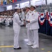 A formal U.S. Navy graduation or awards ceremony taking place in a large indoor facility. A sailor in dress whites is receiving a certificate or document with a handshake from a senior officer, while another officer stands nearby. A large audience of sailors and civilians fills the background, with patriotic red, white, and blue bunting decorations visible.
