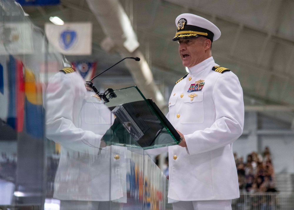 A senior U.S. Navy officer in white dress uniform is delivering a speech at a podium during what appears to be a formal military ceremony. The officer wears multiple service ribbons, gold insignia, and a white combination cover with gold oak leaf scrambled eggs, indicating flag or senior officer rank. An audience is visible in the background, and multiple flags are displayed in the venue.