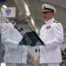A senior U.S. Navy officer in white dress uniform is delivering a speech at a podium during what appears to be a formal military ceremony. The officer wears multiple service ribbons, gold insignia, and a white combination cover with gold oak leaf scrambled eggs, indicating flag or senior officer rank. An audience is visible in the background, and multiple flags are displayed in the venue.