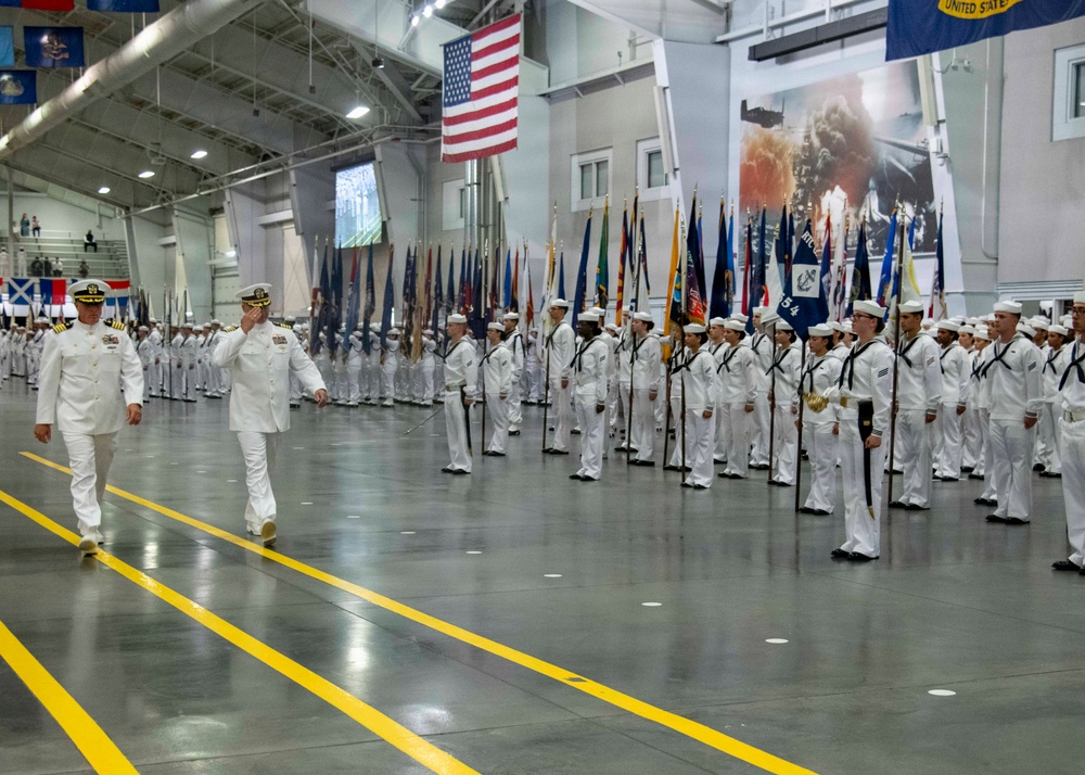 A formal U.S. Navy graduation or pass-in-review ceremony taking place inside a large indoor drill hall. Two senior naval officers in white dress uniforms are walking and rendering salutes while rows of enlisted sailors in white dress uniforms stand at attention. Multiple state and military flags are carried by a color guard formation.