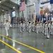 A formal U.S. Navy graduation or pass-in-review ceremony taking place inside a large indoor drill hall. Two senior naval officers in white dress uniforms are walking and rendering salutes while rows of enlisted sailors in white dress uniforms stand at attention. Multiple state and military flags are carried by a color guard formation.