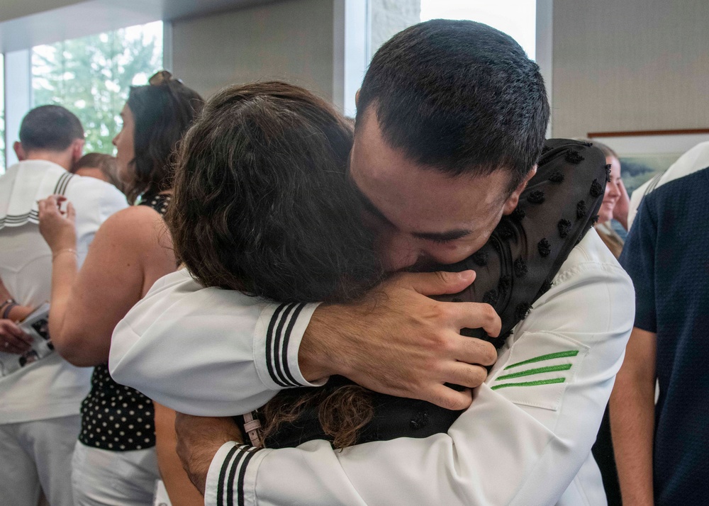 A person in a white U.S. Navy uniform is embracing another individual in a warm, emotional hug in what appears to be an indoor gathering space. The scene suggests a reunion or farewell event, with multiple other people in the background, some also wearing white naval uniforms. The embrace appears heartfelt and emotionally significant.
