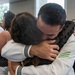 A person in a white U.S. Navy uniform is embracing another individual in a warm, emotional hug in what appears to be an indoor gathering space. The scene suggests a reunion or farewell event, with multiple other people in the background, some also wearing white naval uniforms. The embrace appears heartfelt and emotionally significant.