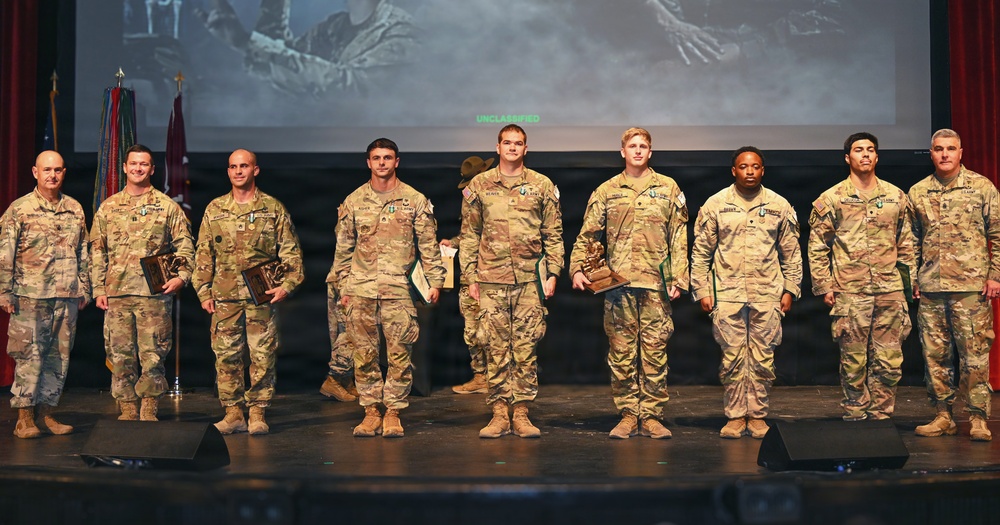 A group of nine U.S. Army soldiers in MultiCam/OCP camouflage uniforms are standing on a stage during what appears to be an awards or recognition ceremony. Several soldiers are holding plaques or trophies, suggesting they are being honored for achievements. A projection screen displaying 'UNCLASSIFIED' text and military flags are visible in the background.
