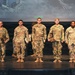 A group of nine U.S. Army soldiers in MultiCam/OCP camouflage uniforms are standing on a stage during what appears to be an awards or recognition ceremony. Several soldiers are holding plaques or trophies, suggesting they are being honored for achievements. A projection screen displaying 'UNCLASSIFIED' text and military flags are visible in the background.