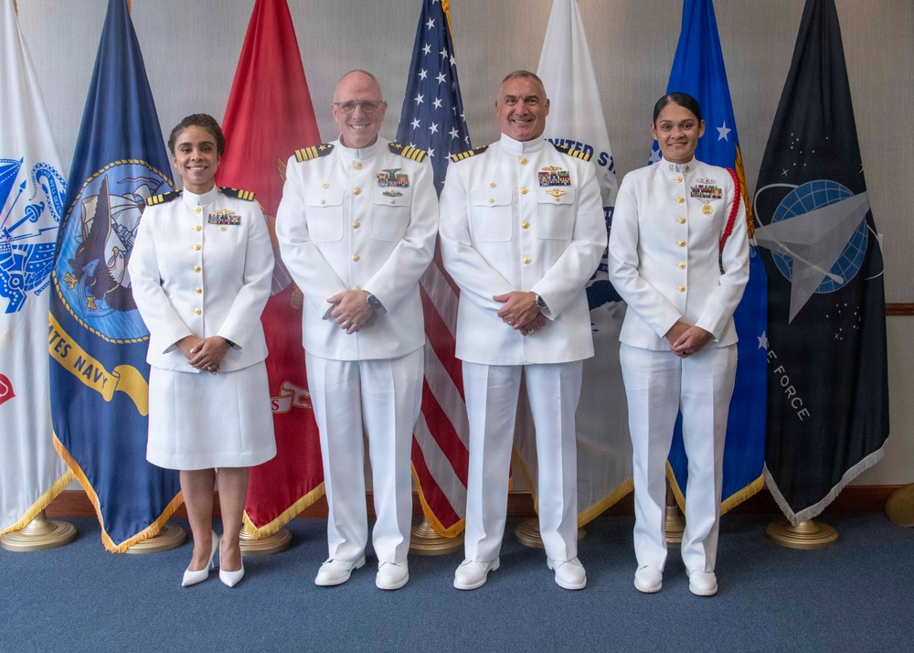 Four U.S. Navy personnel in white dress uniforms are posed for a formal group photograph in front of multiple military service flags. The group consists of two senior officers (center) with more extensive medal ribbons and two junior officers flanking them. The setting appears to be an official military ceremony or event room.