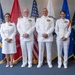 Four U.S. Navy personnel in white dress uniforms are posed for a formal group photograph in front of multiple military service flags. The group consists of two senior officers (center) with more extensive medal ribbons and two junior officers flanking them. The setting appears to be an official military ceremony or event room.